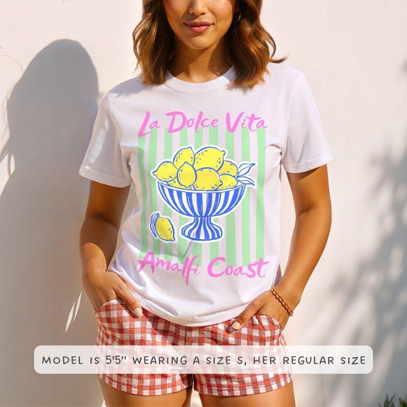 A woman is wearing a White Comfort Colors shirt with a. design featuring a bow of lemons against a striped background and the phrase La Dolce Vita, Amalfi Coast in a playful script font.