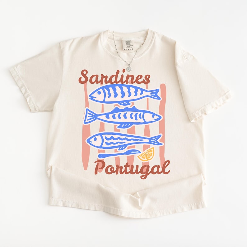 May include: A cream-coloured t-shirt with a graphic of three blue sardines, red vertical stripes, a lemon slice, and the text "Sardines Portugal" in brown. The shirt has a crew neck and short sleeves.