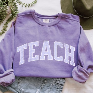 Op de afbeelding: Paarse trui met het woord "TEACH" in witte omrande letters.