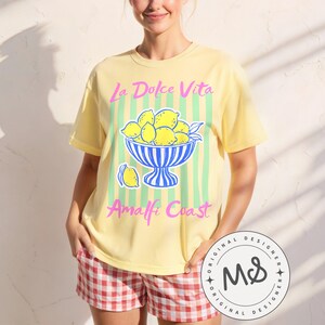 A woman is wearing a Banana Comfort Colors shirt with a. design featuring a bow of lemons against a striped background and the phrase La Dolce Vita, Amalfi Coast in a playful script font.