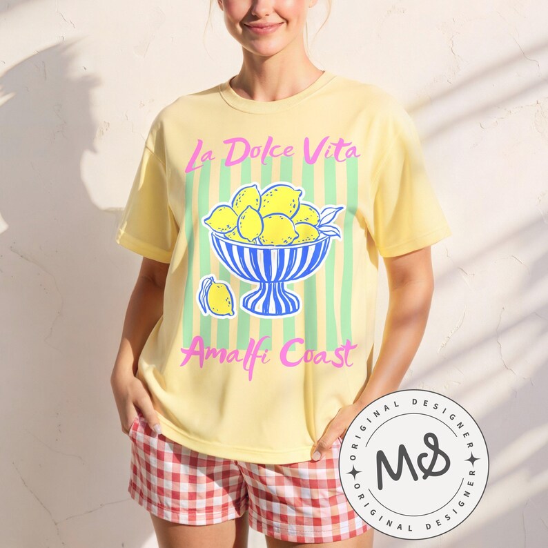 A woman is wearing a Banana Comfort Colors shirt with a. design featuring a bow of lemons against a striped background and the phrase La Dolce Vita, Amalfi Coast in a playful script font.