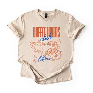 A  flat lay of a Sand  2000 t-shirt with a design featuring a hand-drawn coffee and croissant design that reads Coffee Lover Club, All Day Every Day.