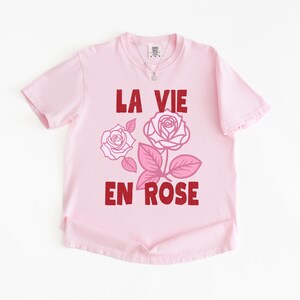 A flat lay of a Blossom pink Comfort Colors t-shirt with bold red lettering that reads “La Vie En Rose.” The graphic features three outlined pink roses with stylized leaves, giving a vintage-inspired romantic look. She pairs the tee with pastel green gingham shorts for a soft, summery aesthetic.