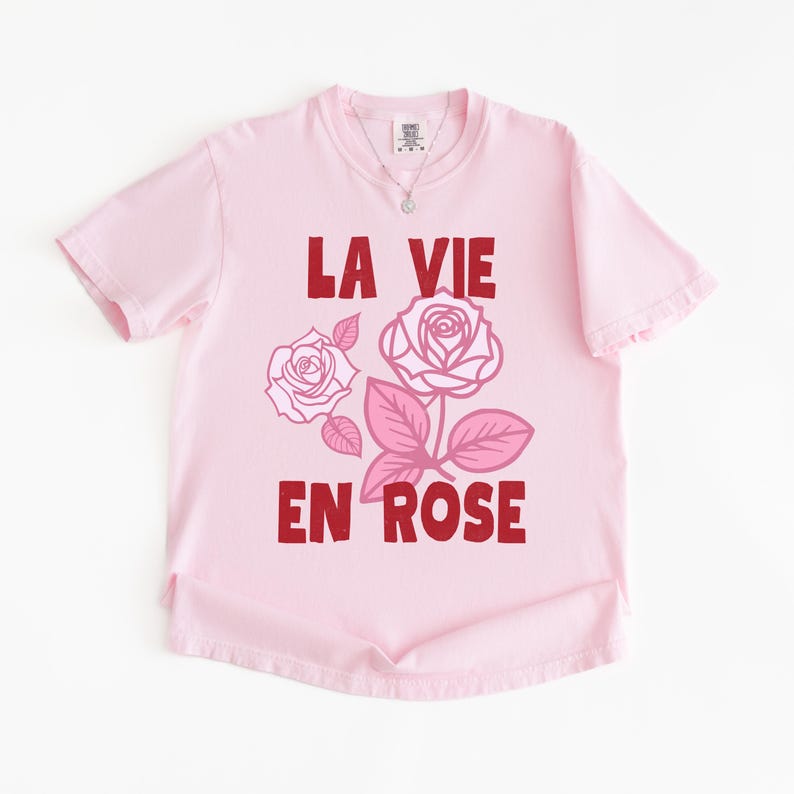 A flat lay of a Blossom pink Comfort Colors t-shirt with bold red lettering that reads “La Vie En Rose.” The graphic features three outlined pink roses with stylized leaves, giving a vintage-inspired romantic look. She pairs the tee with pastel green gingham shorts for a soft, summery aesthetic.