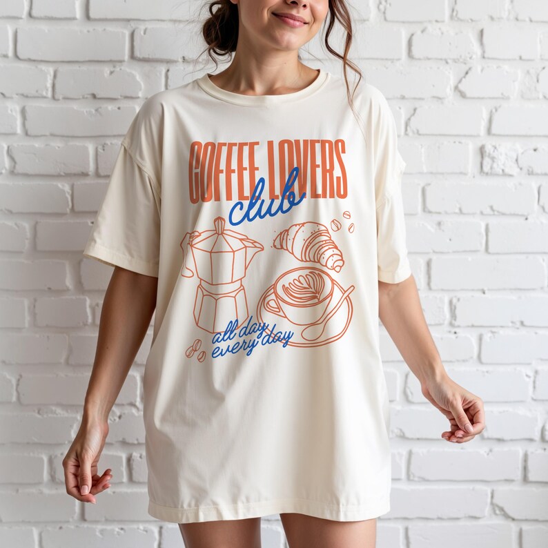 A woman is wearing a Natural Gildan 2000 t-shirt with a design featuring a hand-drawn coffee and croissant design that reads Coffee Lover Club, All Day Every Day.