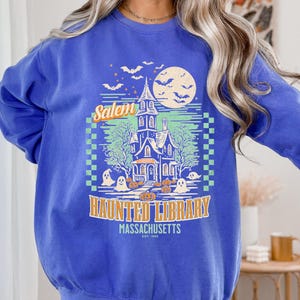 A woman is wearing an oversized Flo BlueComfort Colors sweatshirt with a design featuring a haunted library with ghosts and pumpkins and the phrase Salem Haunted Library, Massachusetts.