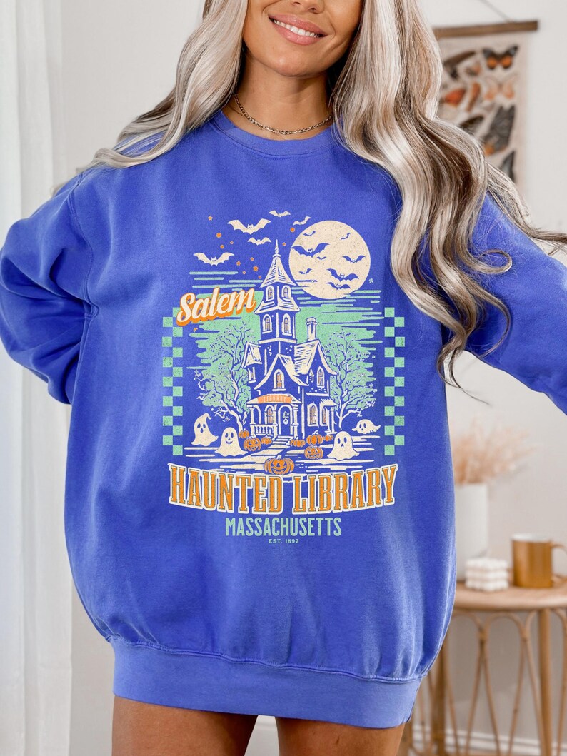 A woman is wearing an oversized Flo BlueComfort Colors sweatshirt with a design featuring a haunted library with ghosts and pumpkins and the phrase Salem Haunted Library, Massachusetts.
