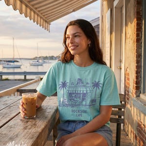 Peut inclure: Un t-shirt bleu clair avec un dessin de caf&eacute;, des palmiers et le texte "Dockside Cafe". L'image montre une personne assise &agrave; une table en bois avec un verre de caf&eacute; glac&eacute;, avec des voiliers en arri&egrave;re-plan.