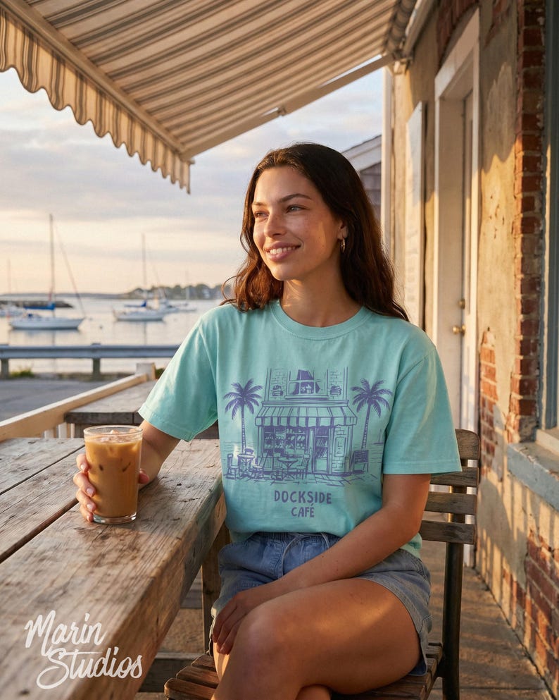Peut inclure: Un t-shirt bleu clair avec un dessin de caf&eacute;, des palmiers et le texte "Dockside Cafe". L'image montre une personne assise &agrave; une table en bois avec un verre de caf&eacute; glac&eacute;, avec des voiliers en arri&egrave;re-plan.