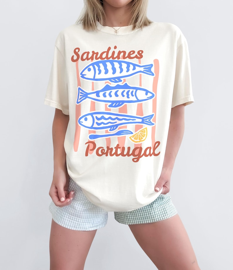 A woman is wearing an Ivory Comfort Colors shirt with a design featuring three sardines in a hand-drawn style and the phrase Sardines, Portugal in a script style font.