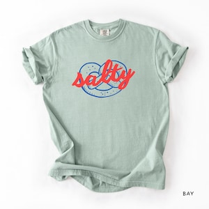A flat lay of a Bay Comfort Colors shirt with a design featuring a pretzel and the phrase Salty over the top of it.