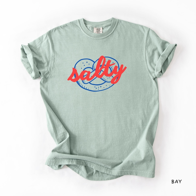 A flat lay of a Bay Comfort Colors shirt with a design featuring a pretzel and the phrase Salty over the top of it.