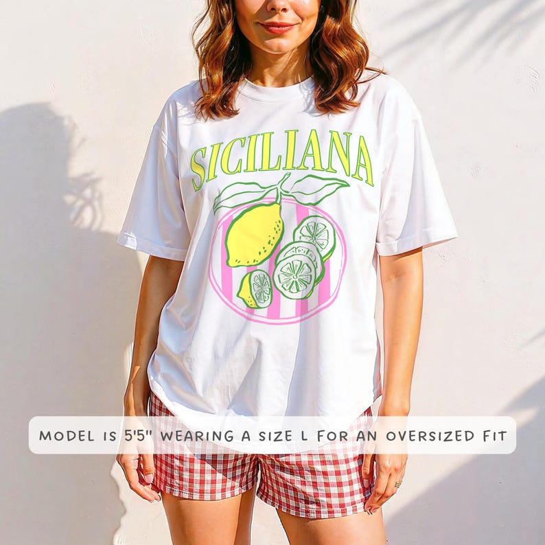 A woman is wearing a white Comfort Colors shirt with a design featuring a hand-drawn lemon and lemon slices on a plate, and the phrase Siciliana at the top in a serif font.