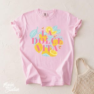 May include: A light pink t-shirt with the words "LA DOLCE VITA" in pink and yellow, surrounded by a lemon and leaf design. The shirt has rolled-up sleeves and a relaxed fit. A beige tote bag is in the background.