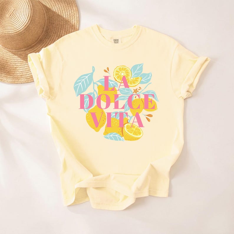 May include: A light yellow t-shirt featuring a graphic of lemons, leaves, and the text "LA DOLCE VITA" in pink and yellow. The design includes lemons and leaves in a vibrant, summery style.