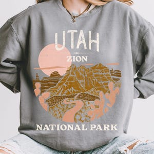 Woman wearing a Grey oversized sweatshirt with a vintage graphic of Zion National Park in Utah, featuring canyons, cacti, and a sunset design, styled with ripped jeans.