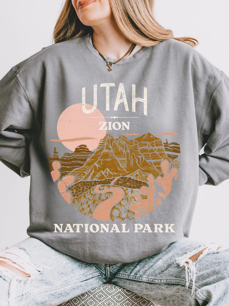 Woman wearing a Grey oversized sweatshirt with a vintage graphic of Zion National Park in Utah, featuring canyons, cacti, and a sunset design, styled with ripped jeans.