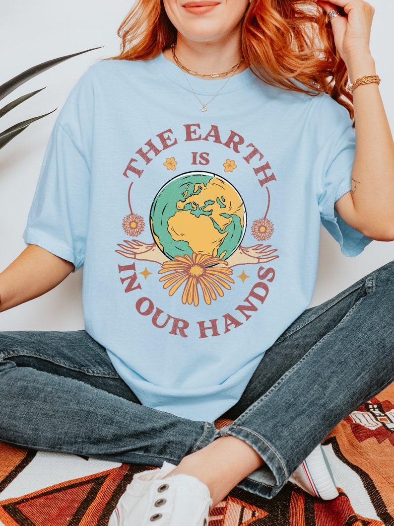 A woman is wearing a chambray blue Comfort Colors shirt with an Earth Day design that says The Earth is in Your Hands.