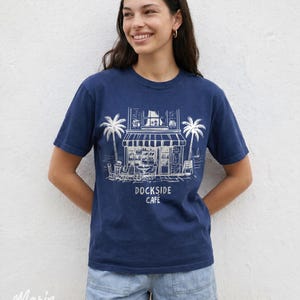 Peut inclure: T-shirt bleu marine avec une illustration blanche d'un caf&eacute;, de palmiers et le texte "Dockside Cafe". Le t-shirt est en mati&egrave;re souple et convient aux tenues d&eacute;contract&eacute;es. Le logo "Marin Studios" est visible.