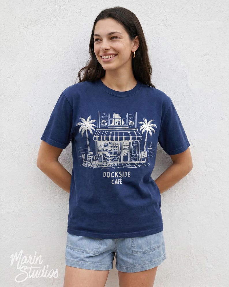 Peut inclure: T-shirt bleu marine avec une illustration blanche d'un caf&eacute;, de palmiers et le texte "Dockside Cafe". Le t-shirt est en mati&egrave;re souple et convient aux tenues d&eacute;contract&eacute;es. Le logo "Marin Studios" est visible.