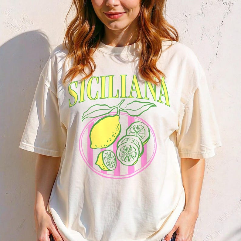A woman is wearing an ivory Comfort Colors shirt with a design featuring a hand-drawn lemon and lemon slices on a plate, and the phrase Siciliana at the top in a serif font.