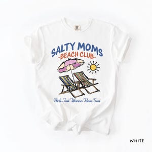 A White Comfort Colors shirt ona simple white background with a desgn  featuring a colorful hand-drawn scene of a striped beach umbrella and two chairs with the phrase Salty Moms Beach Club at the top and Girls Just Wanna Have Fun at the bottom of the design.
