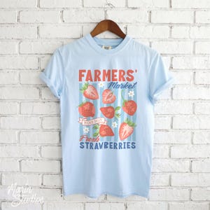 May include: Light blue t-shirt with a graphic featuring the words "FARMERS' Market" and "Fresh Strawberries." The design includes illustrations of strawberries and flowers. The shirt is hanging on a wooden hanger.