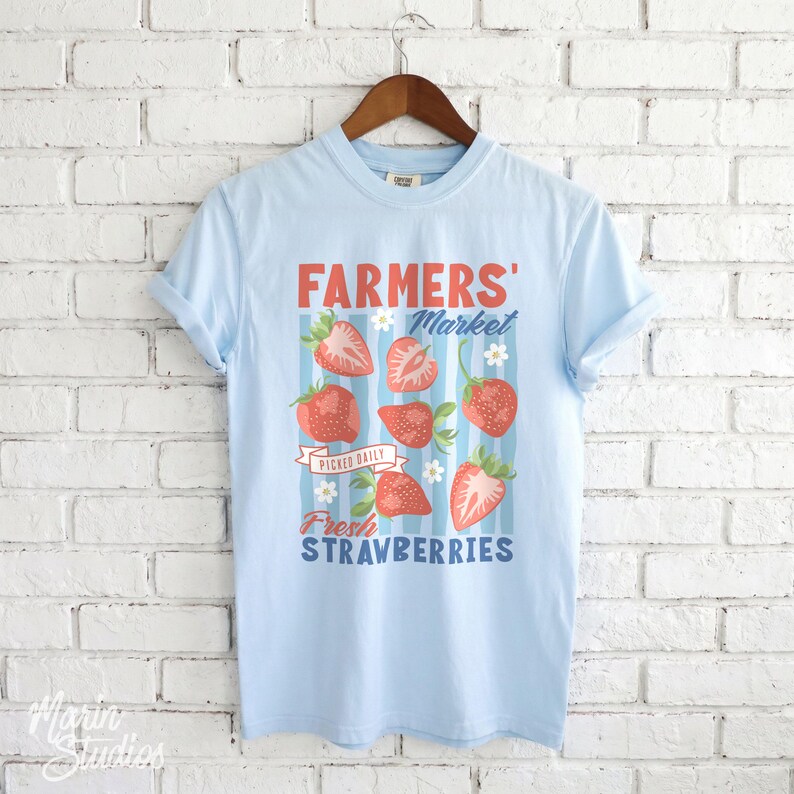 May include: Light blue t-shirt with a graphic featuring the words "FARMERS' Market" and "Fresh Strawberries." The design includes illustrations of strawberries and flowers. The shirt is hanging on a wooden hanger.