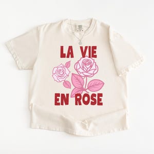 A flat lay of an Ivory Comfort Colors t-shirt with bold red lettering that reads “La Vie En Rose.” The graphic features three outlined pink roses with stylized leaves, giving a vintage-inspired romantic look. She pairs the tee with pastel green gingham shorts for a soft, summery aesthetic.