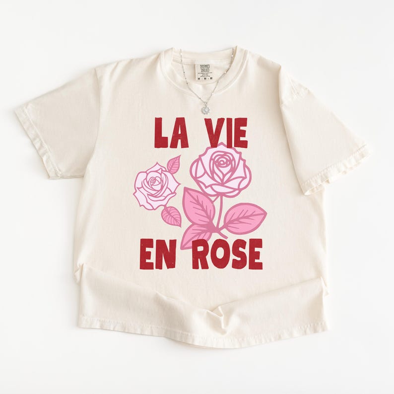 A flat lay of an Ivory Comfort Colors t-shirt with bold red lettering that reads “La Vie En Rose.” The graphic features three outlined pink roses with stylized leaves, giving a vintage-inspired romantic look. She pairs the tee with pastel green gingham shorts for a soft, summery aesthetic.