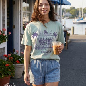 Peut inclure: Une personne porte un t-shirt vert clair avec un graphisme violet d'un caf&eacute; et le texte "DOCKSIDE CAFE". Elle porte &eacute;galement un short en jean bleu et tient une tasse en plastique transparente avec du caf&eacute; glac&eacute;. L'arri&egrave;re-plan comprend un b&acirc;timent et un port.
