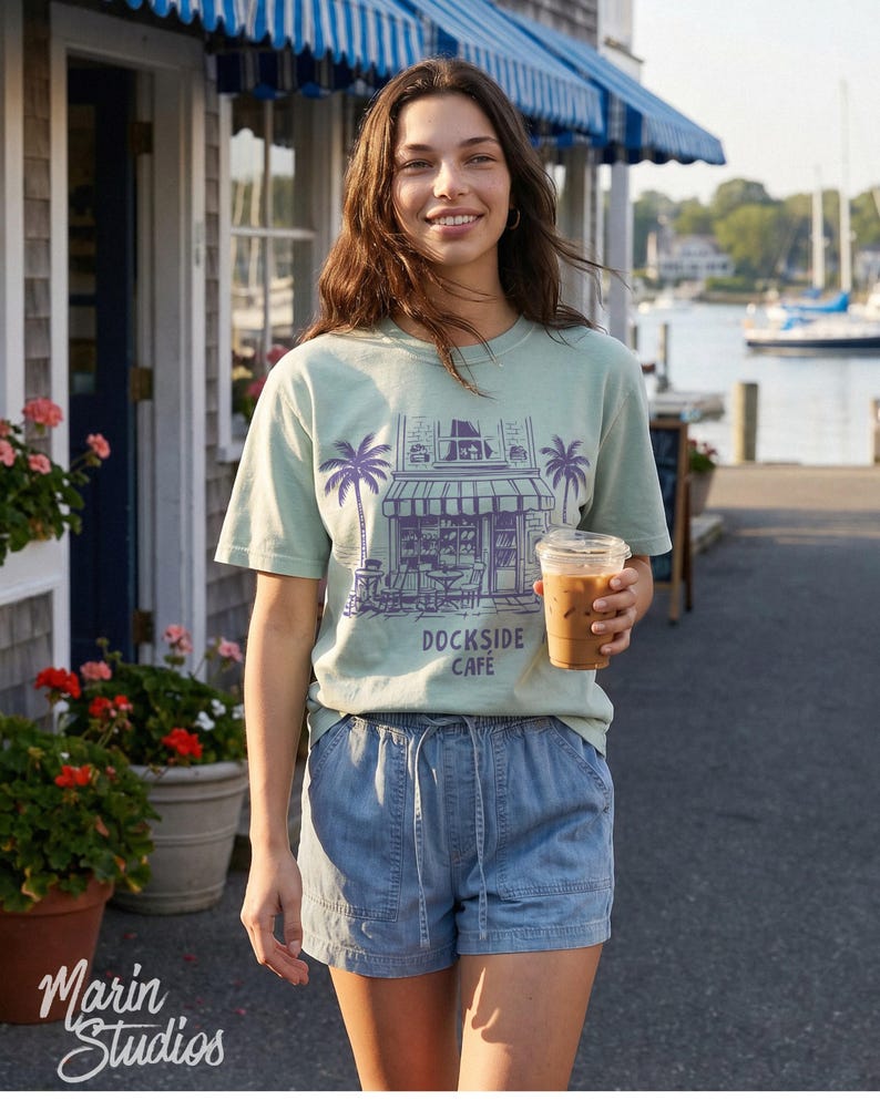 Peut inclure: Une personne porte un t-shirt vert clair avec un graphisme violet d'un caf&eacute; et le texte "DOCKSIDE CAFE". Elle porte &eacute;galement un short en jean bleu et tient une tasse en plastique transparente avec du caf&eacute; glac&eacute;. L'arri&egrave;re-plan comprend un b&acirc;timent et un port.