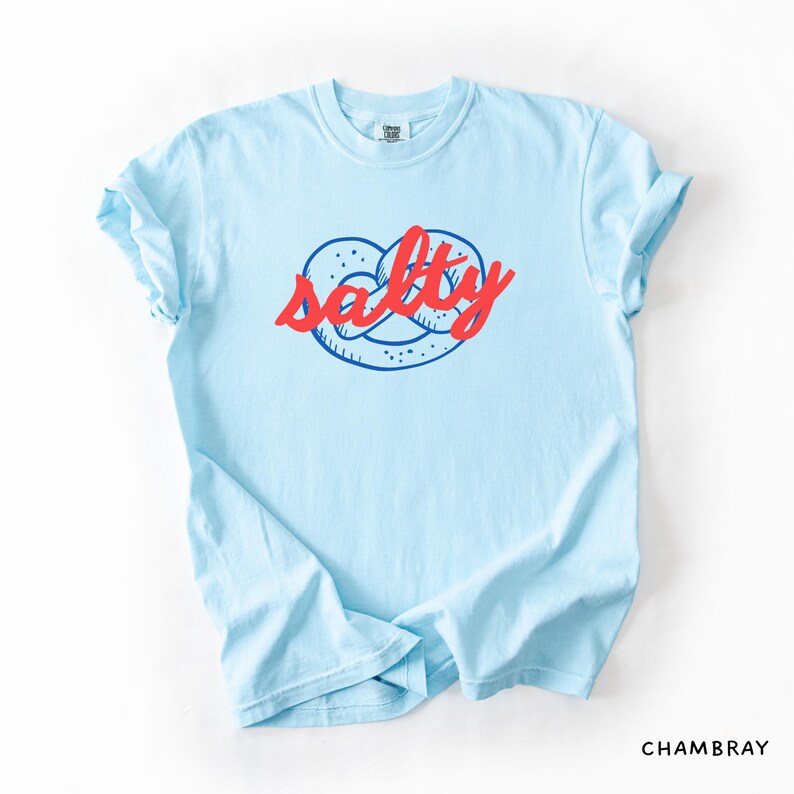 A flat lay of a Chambray Comfort Colors shirt with a design featuring a pretzel and the phrase Salty over the top of it.