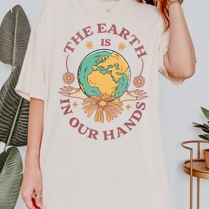 A woman is wearing an ivory Comfort Colors shirt with an Earth Day design that says The Earth is in Your Hands.