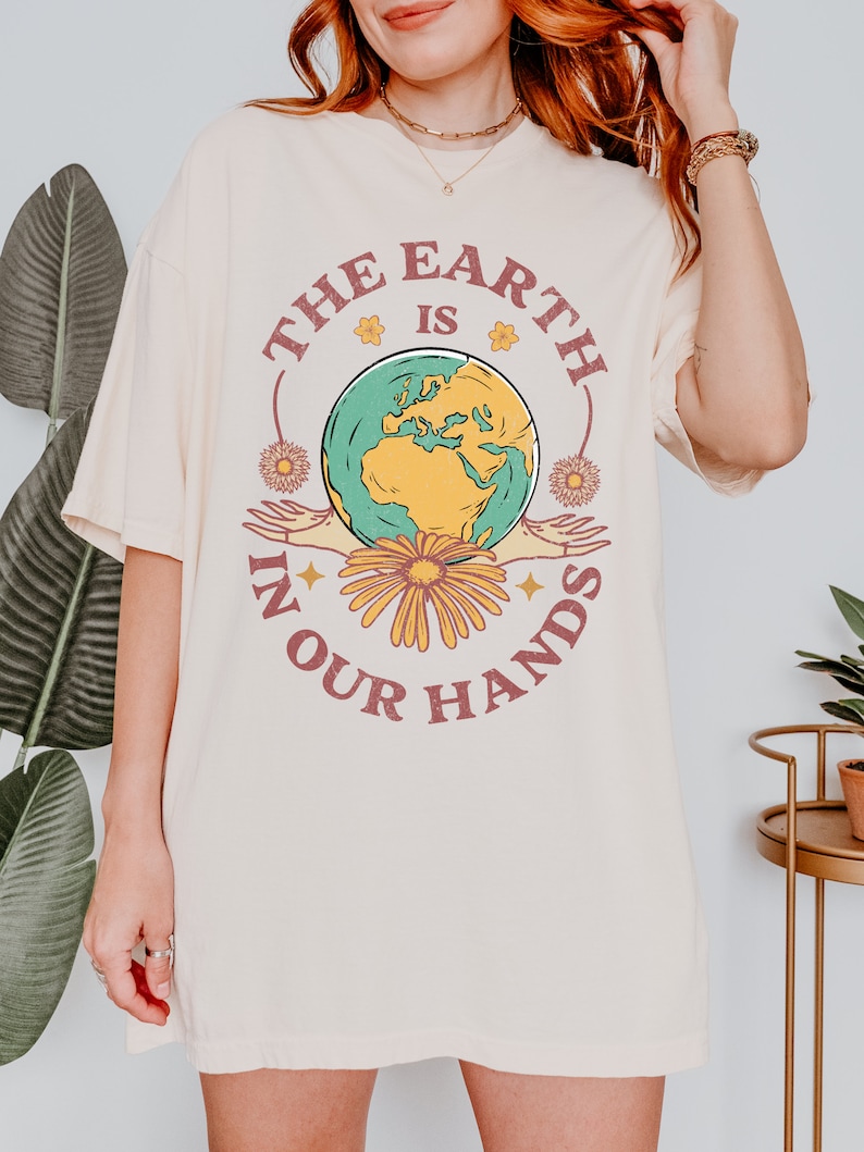 A woman is wearing an ivory Comfort Colors shirt with an Earth Day design that says The Earth is in Your Hands.