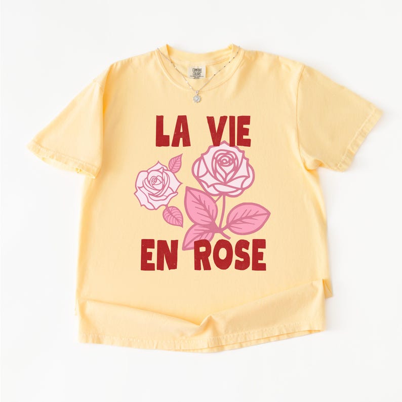 A flat lay of a Butter yellow Comfort Colors t-shirt with bold red lettering that reads “La Vie En Rose.” The graphic features three outlined pink roses with stylized leaves, giving a vintage-inspired romantic look. She pairs the tee with pastel green gingham shorts for a soft, summery aesthetic.