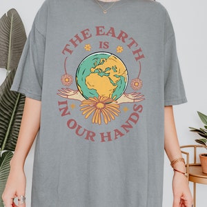 A woman is wearing a grey Comfort Colors shirt with an Earth Day design that says The Earth is in Your Hands.