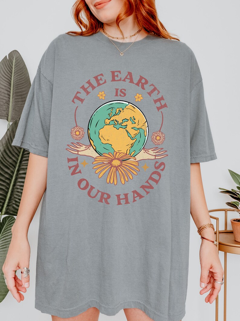 A woman is wearing a grey Comfort Colors shirt with an Earth Day design that says The Earth is in Your Hands.
