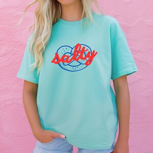 A woman is wearing a Chalky Mint Comfort Colors shirt with a design featuring a pretzel and the phrase Salty over the top of it.