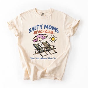An Ivory Comfort Colors shirt ona simple white background with a desgn  featuring a colorful hand-drawn scene of a striped beach umbrella and two chairs with the phrase Salty Moms Beach Club at the top and Girls Just Wanna Have Fun at the bottom of the design.