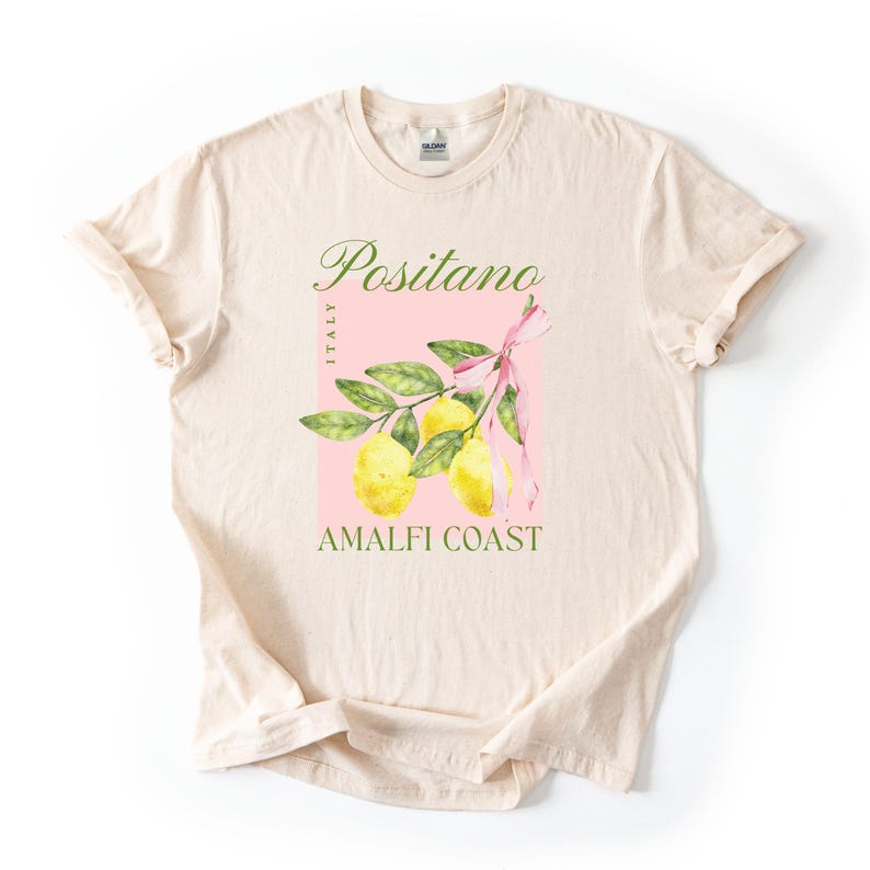 A flat lay of a Natural Gildan 2000 unisex T-shirt with a design featuring a branch of lemons with a long pink coquette bow tied around it and the phrase Positano, Amalfi Coast, Italy.