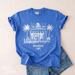 Peut inclure: Un t-shirt bleu avec une illustration blanche d'un caf&eacute;, de palmiers et le texte "DOCKSIDE CAF&Eacute;". Le t-shirt est associ&eacute; &agrave; un short en jean. Le design &eacute;voque un th&egrave;me c&ocirc;tier.