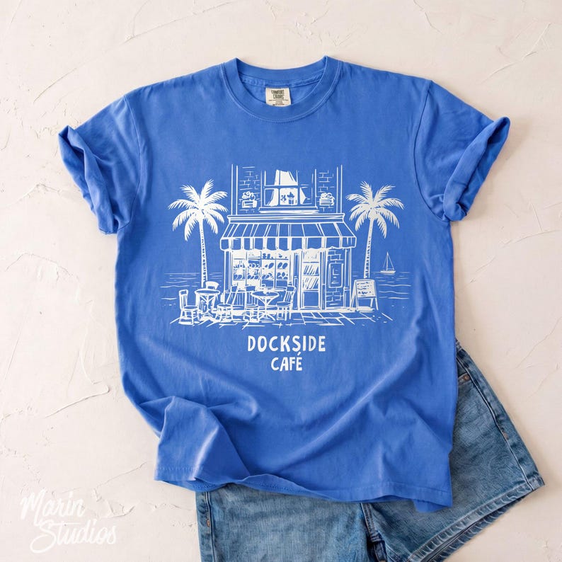 Peut inclure: Un t-shirt bleu avec une illustration blanche d'un caf&eacute;, de palmiers et le texte "DOCKSIDE CAF&Eacute;". Le t-shirt est associ&eacute; &agrave; un short en jean. Le design &eacute;voque un th&egrave;me c&ocirc;tier.