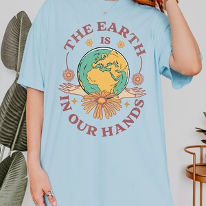 A woman is wearing a chambray blue Comfort Colors shirt with an Earth Day design that says The Earth is in Your Hands.