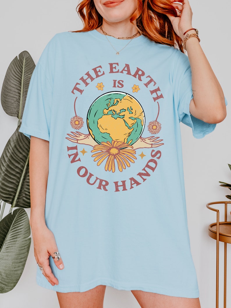 A woman is wearing a chambray blue Comfort Colors shirt with an Earth Day design that says The Earth is in Your Hands.