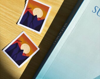 May include: Two sticker designs featuring a mountain range silhouette with a sunset and a full moon. The stickers are on a wooden surface next to a blue book with a wave pattern.
