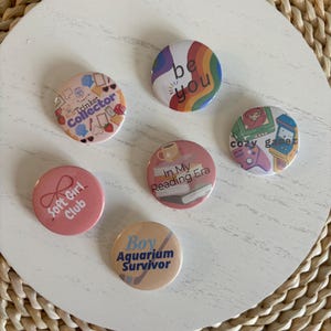 May include: Seven circular pin badges with different designs and text, including "be you", "Soft Girl Club", and "Boy Aquarium Survivor". The badges feature pink, blue, and rainbow colours. They are arranged on a white surface, with a woven background.