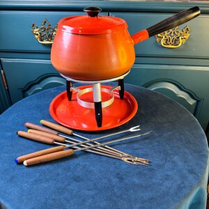 May include: A bright orange fondue pot with a matching lid, a long black handle, and a black knob. The pot rests on a white metal stand with black feet and a circular orange base. Six metal fondue forks with wooden handles are included.