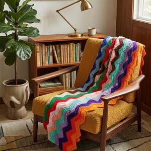May include: A crochet blanket featuring a vibrant chevron pattern in horizontal stripes of orange, purple, light blue, white, red, green, and brown. The blanket is draped over a mustard yellow textured armchair with wooden armrests and legs.