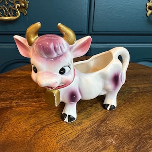 May include: A ceramic cow creamer with a white body, pink and purple accents, and gold horns. The cow has a red collar and a gold bell. The creamer is designed with a handle and a spout for pouring.